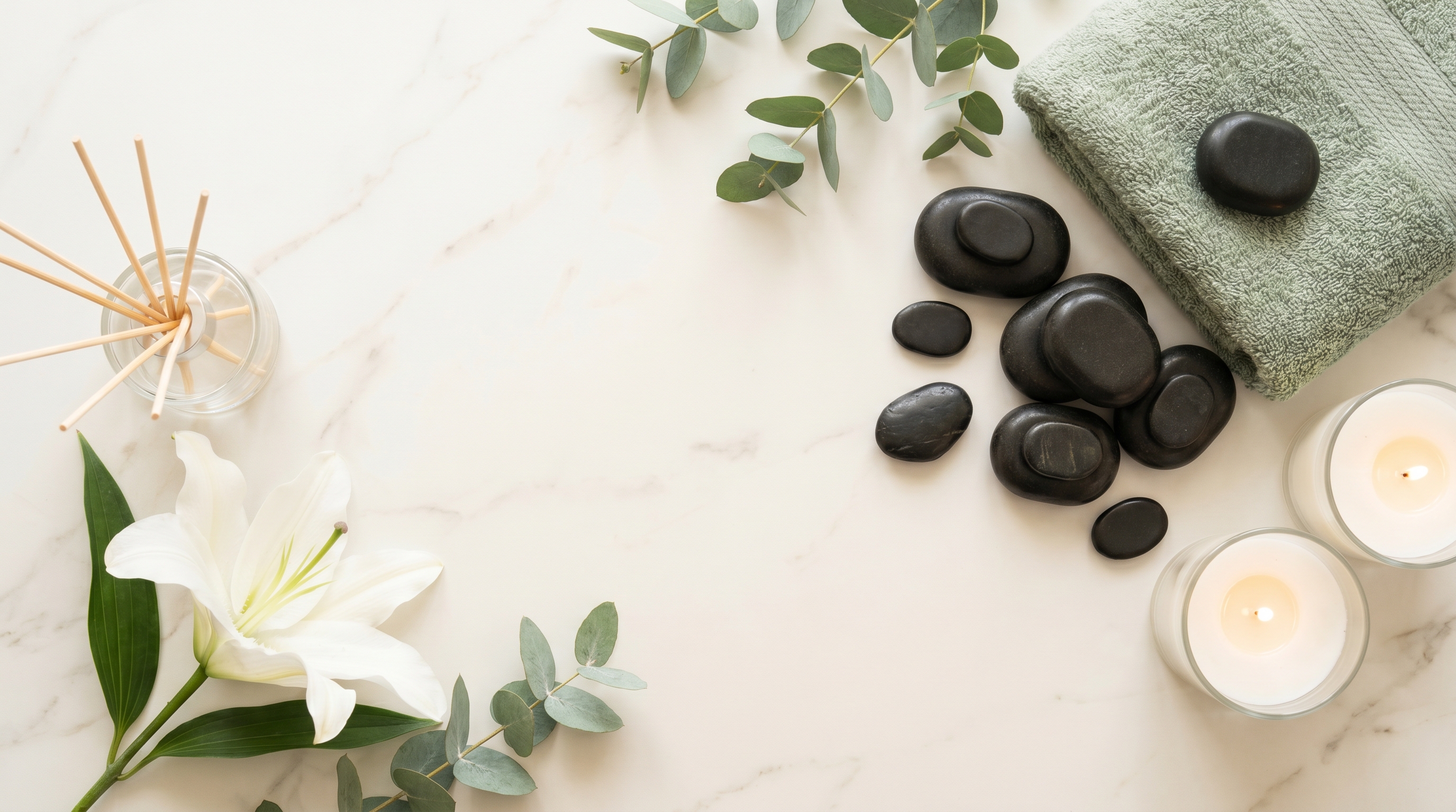A calm spa flatlay with massage stones, a sage towel, candles, a white lily and eucalyptus